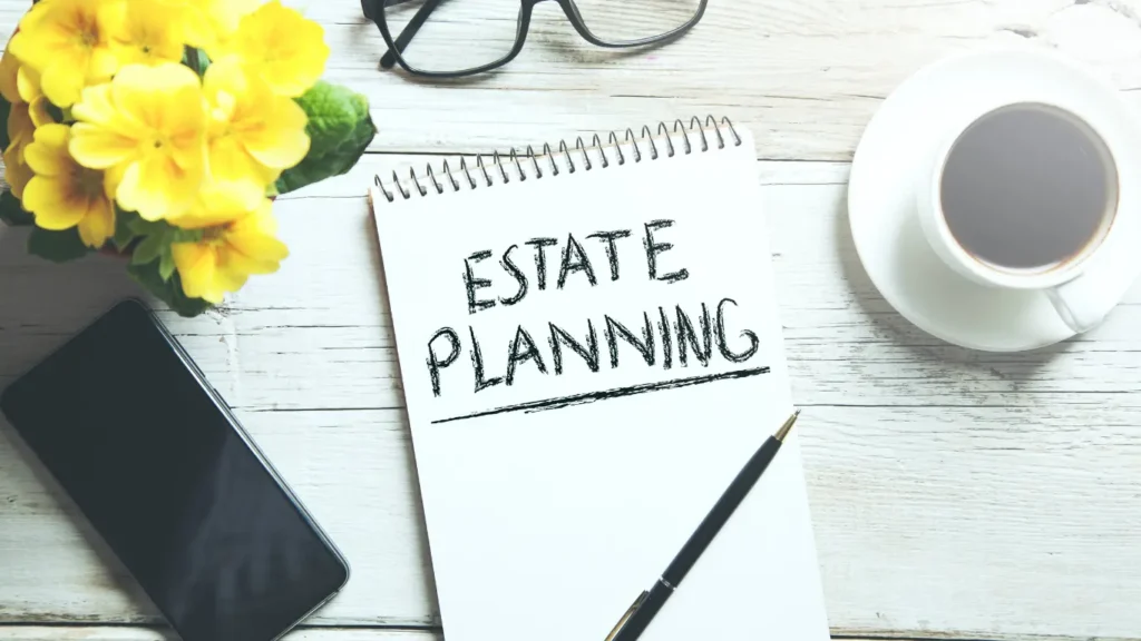 Estate planning concept with a notebook labeled “Estate Planning,” a pen, smartphone, eyeglasses, flowers, and a cup of coffee on a desk.